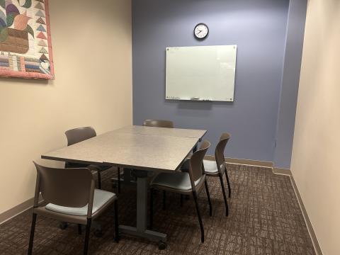 Meeting room with two tables pushed together and 5 chairs around them. An erasable whiteboard and a clock are on a blue wall. A corner of a quilted wall hanging is visible on a beige wall.